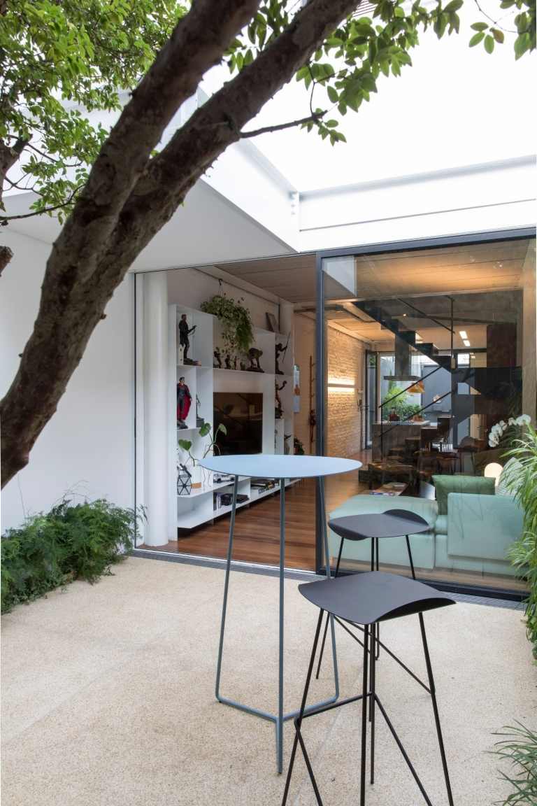 شجرة في الفناء مع طاولة عالية التصميم وكراسي بار تطل على غرفة المعيشة بجدران من الطوب الأبيض ووحدة جدارية